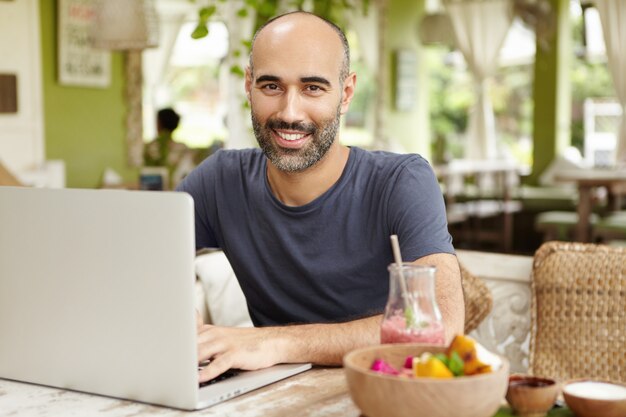 Bearded businessman dressed casually checking email on his laptop during breakfast, sitting at nice cafe, drinking smoothie with happy confident face expression, enjoying vacations