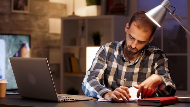 Bearded businessman sitting at desk in home office during night hours and signing documents.