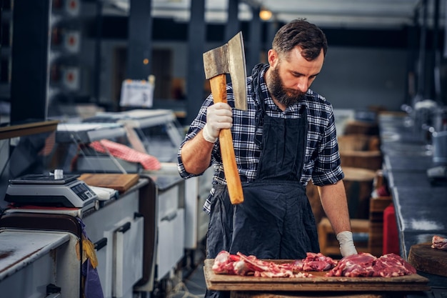 Bearded butcher dressed in a fleece shirt cuts meat with a bloody axe.