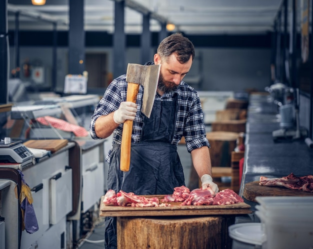 Foto gratuita il macellaio barbuto vestito con una camicia di pile taglia la carne con un'ascia insanguinata.