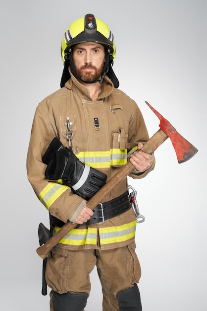 Bearded caucasian firefighter in full gear holding red axe for fire fighting in studio front view of