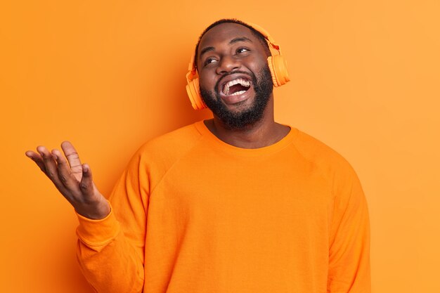 Bearded dark skinned man raises hand laughs positively listens audio track via stereo headphones