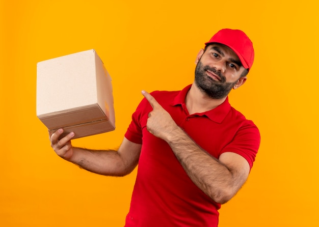 Bearded delivery man in red uniform and cap holding box package pointing with finger to it smiling confident standing over orange wall