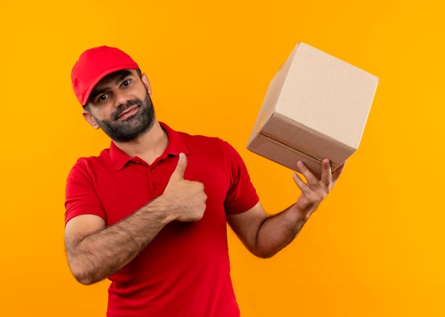 Uomo barbuto di consegna in uniforme rossa e cappuccio che tiene il pacchetto della scatola che mostra i pollici in su in piedi positivo e felice sopra la parete arancione