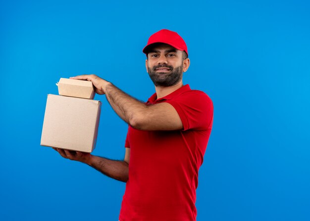 파란색 벽 위에 서 자신감 미소로 상자 패키지를 들고 빨간 유니폼과 모자 수염 배달 남자