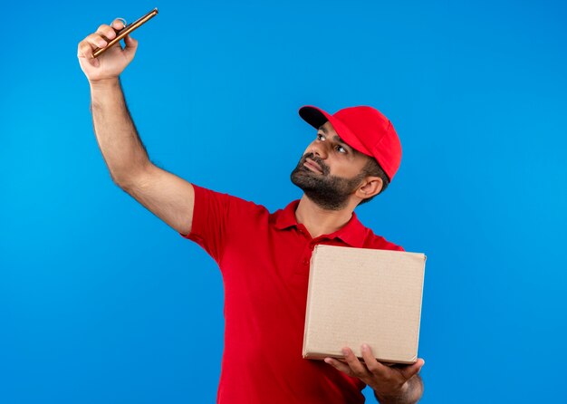 Бородатый курьер в красной форме и кепке, держащий коробку, делает селфи на своем смартфоне, стоящем над синей стеной