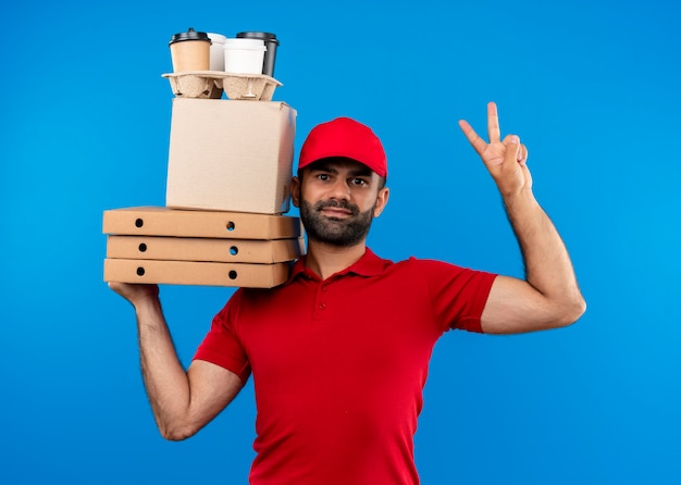 Bearded delivery man in red uniform and cap holding cardboard boxes looking aside with smile on face showing victory sign standing over blue wall