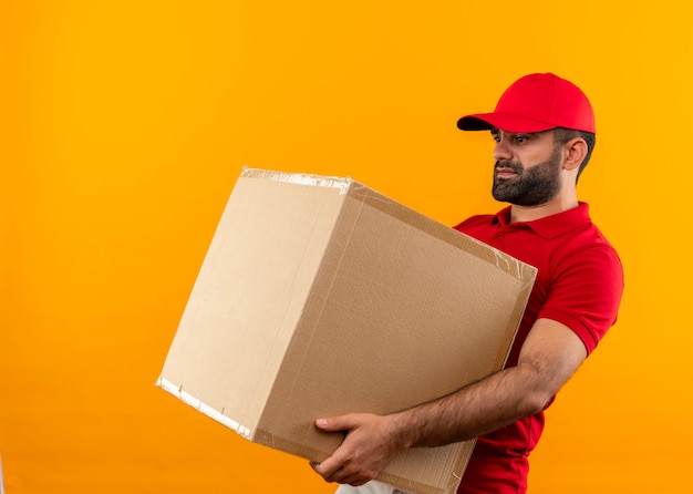 Bearded delivery man in red uniform and cap holding large box package suffering from heavy weight standing over orange wall