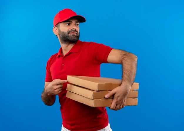 Bearded delivery man in red uniform and cap holding stack of pizza boxes looking aside pointing with finger at boxes standing over blue wall