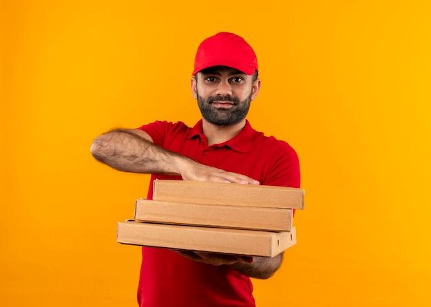 Bearded delivery man in red uniform and cap holding stack of pizza boxes offering with friendly smile standing over orange wall