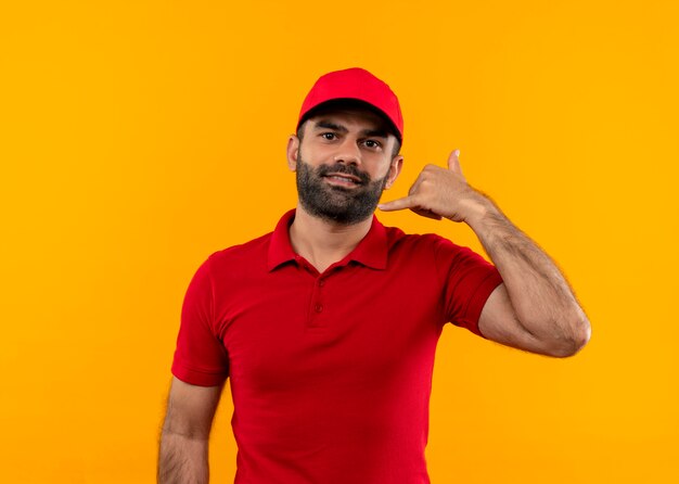 Bearded delivery man in red uniform and cap looking confident making call me gesture standing over orange wall