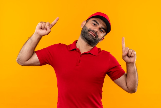 주황색 벽 위에 서있는 미소로 검지 손가락으로 가리키는 빨간 유니폼과 모자 수염 배달 남자
