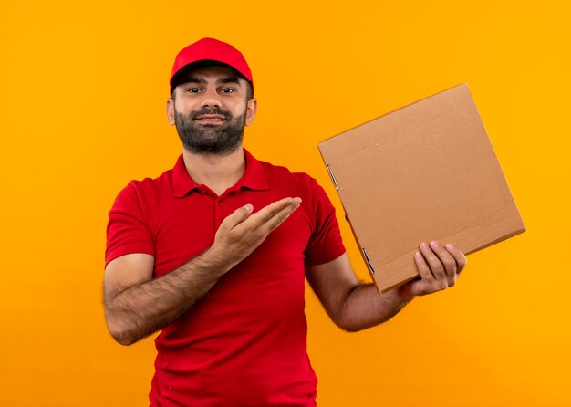빨간 제복을 입은 수염 배달 남자와 그의 손의 팔로 피자 상자를 제시하는 모자는 오렌지 벽 위에 자신감이 서 웃고