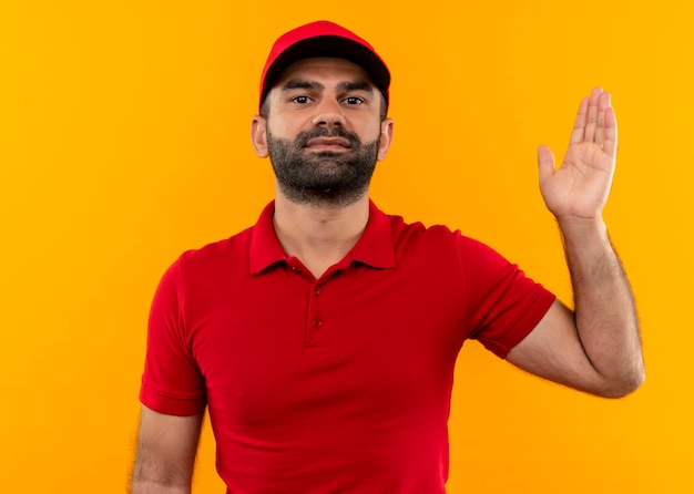 Bearded delivery man in red uniform and cap raising arm looking confident standing over orange wall