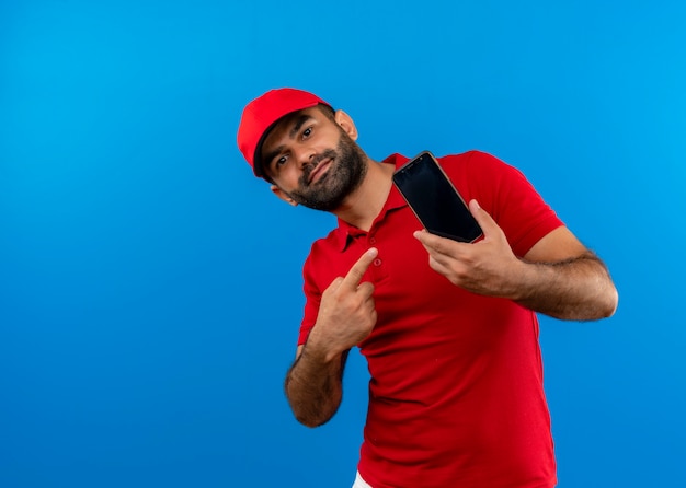Бородатый курьер в красной форме и кепке показывает свой смартфон, указывая пальцем на него, улыбаясь, стоя у синей стены