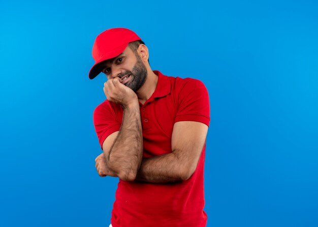 Bearded delivery man in red uniform and cap stressed and nervous biting nails standing over blue wall