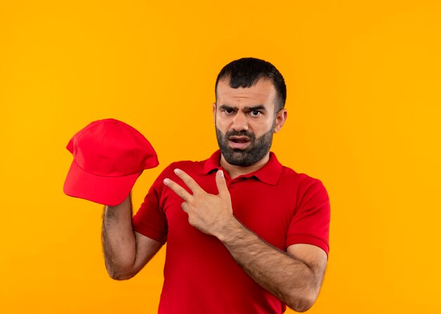 Bearded delivery man in red uniform holding cap pointing with finger to it looking confused and displeased standing over orange wall