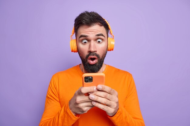 ひげを生やした感情的な男がスマートフォンのディスプレイを見つめる携帯電話アプリを使用してスマートフォンの凝視を印象づけ、音楽を聴くためにワイヤレスヘッドフォンを使用する