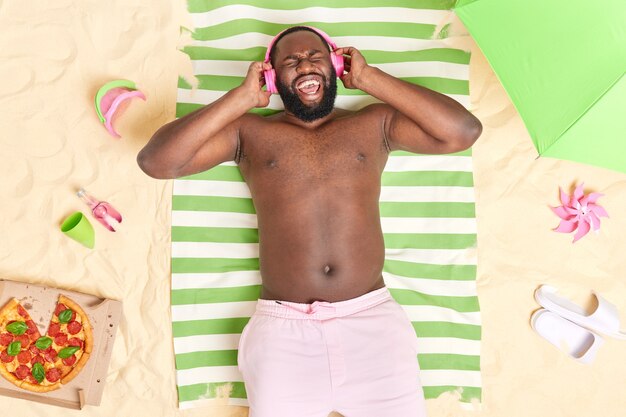 Bearded ethnic guy has upbeat mood listens music via wireless headphones wears shorts poses on green striped towel at sandy beach has lazy day enjoys summer time