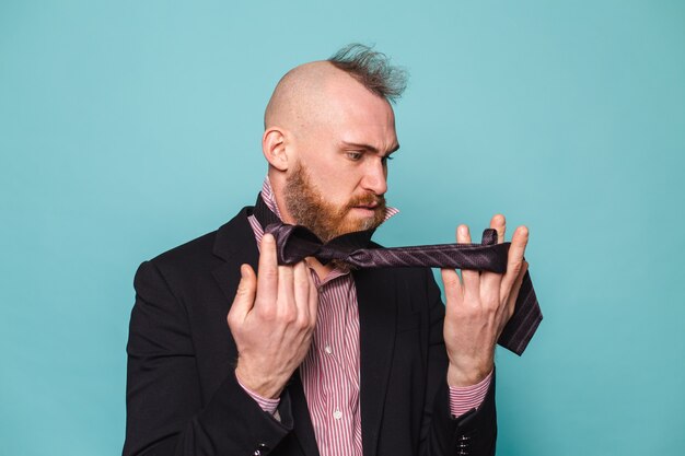 Bearded european businessman in dark suit isolated, angry trying to tie a tie dont know how to do it