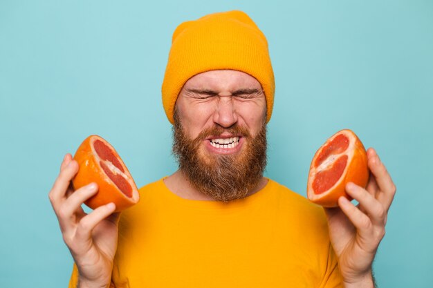 Bearded european man in yellow shirt isolated, holding grapefruit tastes, wrinkles from bitterness