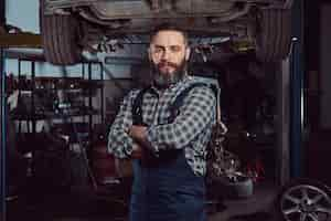 Free photo bearded expert mechanic dressed in a uniform, standing with crossed arms against a car on a lift in the garage.