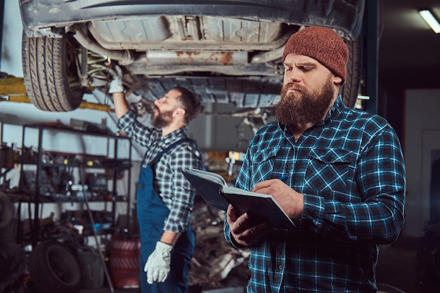 Bearded expert mechanic records results of inspecting a car while his partner checking a problem with a car suspension. A scene from a service station.