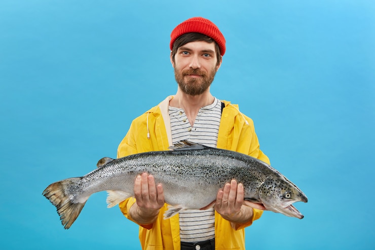 Free Photo bearded fisherman in yellow anorak and red hat holding huge fish in hands, demonstrating his successful catch. horizontal portrait of skilled workman posing with big salmon on blue wall