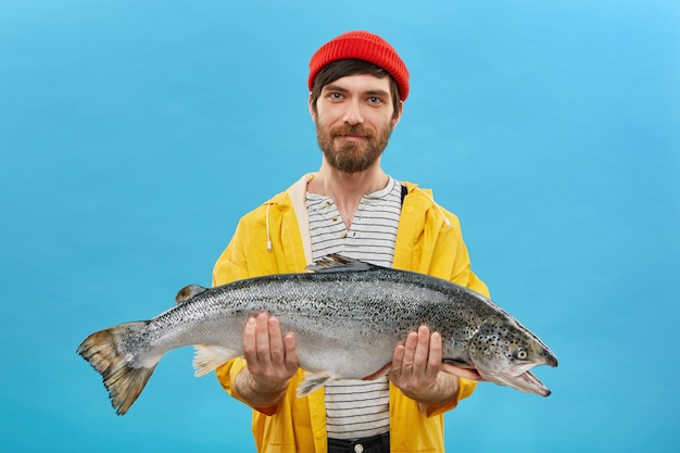 Бородатый рыбак в желтом куртке и красной шляпе держит в руках огромную рыбу, демонстрируя свой успешный улов. Горизонтальный портрет квалифицированного рабочего, позирующего с большим лососем на синей стене