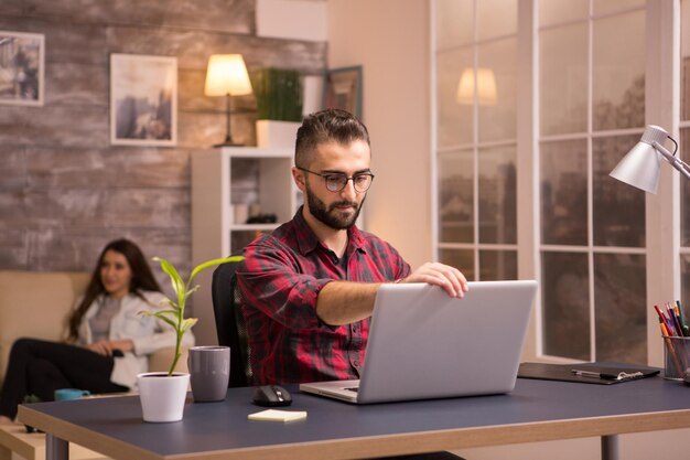 Foto gratuita libero professionista barbuto che apre il suo laptop per iniziare a lavorare sul soggiorno. fidanzata rilassante sul divano a guardare la tv in background.