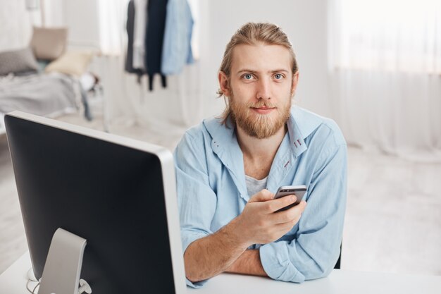 Bearded good-looking male office worker with gentle smile reads notification on smart phone, sits in front of screen at coworking space with cell phone, sends feedback to coworkers, browses internet