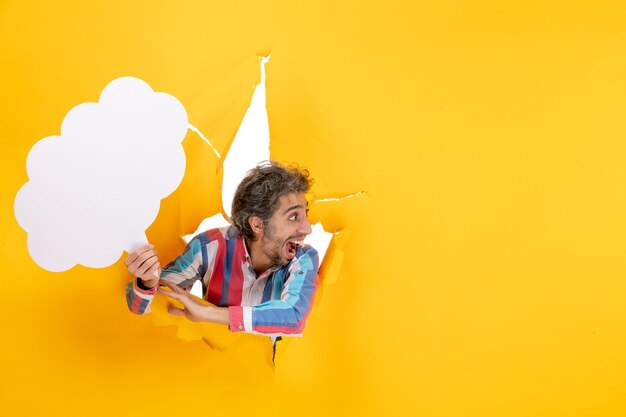 Bearded guy holding white cloud-shaped paper and looking at something with surprised facial expression in a torn hole and free background in yellow paper