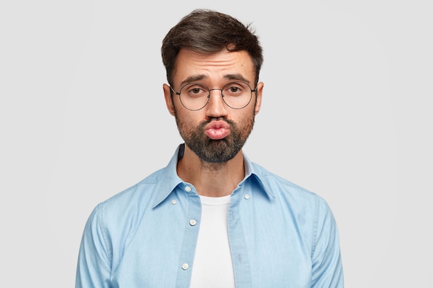 Bearded guy makes grimace, keeps mouth round, has thick stubble, dressed in fashionable stylish shirt