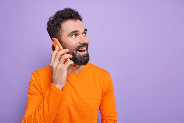 Bearded guy with cheerful expression expresses sinecere emotions talks via smartphone looks away has happy conversation dressed in orange long sleeved jumper 