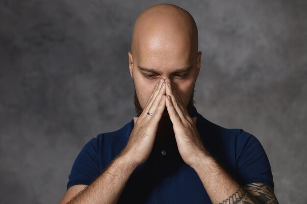 Bearded guy with shaved head having cold, holding hands at his nose as if going to sneeze. Bald male feeling depressed covering face, pondering, searching for solution to problem. Body language