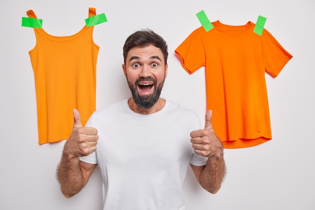 Bearded handsome man keeps thumb up recommends something has positive expression dressed in casual clothes poses against white wall with t shirt and shirt plastered