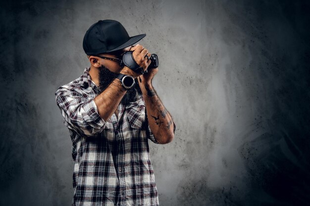 A bearded hipster amateur photographer with tattoos on arms, dressed in a fleece shirt holds compact DSLR camera over grey background.