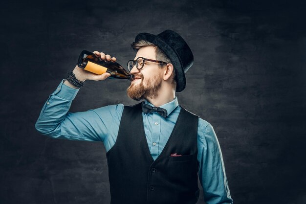 A bearded hipster male dressed in a blue shirt, elegant waistcoat and top hat drinks craft bottle beer.