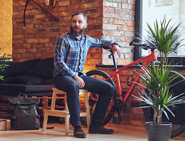 A bearded hipster male dressed in a fleece shirt sits on a wooden chair, near the window in a room with loft interior