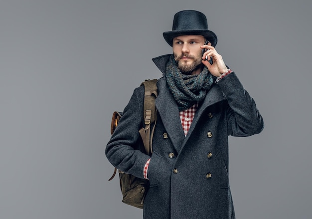 A bearded hipster male dressed in a gray jacket and a cylinder hat isolated on grey background.