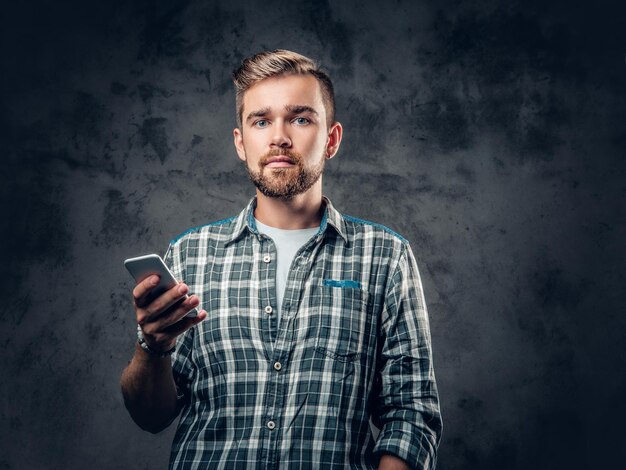 Бородатый мужчина-хипстер в флисовой рубашке держит смартфон на сером фоне.