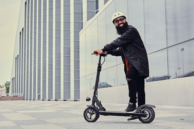 Bearded hipster male vaping and riding by electric scooter over modern building.