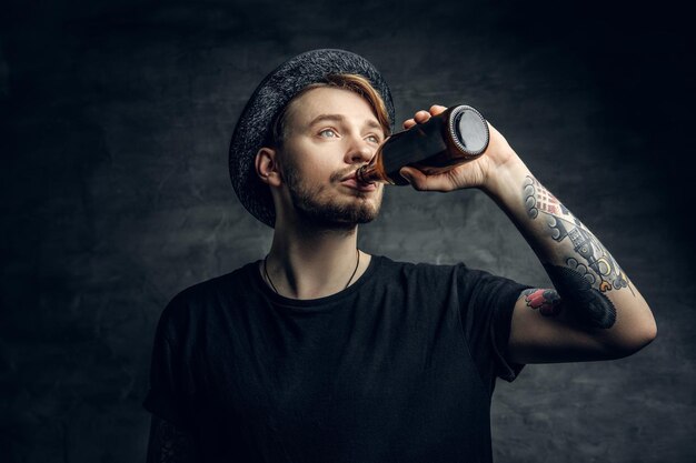 Bearded hipster male with tattoos on his arms, dressed in a black t shirt and top hat drinks craft bottled beer.