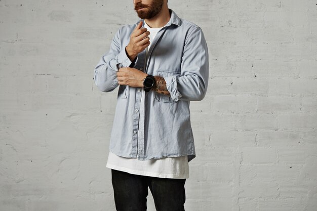 Bearded hipster unbuttoning and rolling up the second sleeve of his casual faded blue denim shirt with white walls