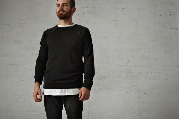 Bearded hipster wearing a blank black longsleeve shirt with a white t-shirt underneath and black jeans on white brick wall