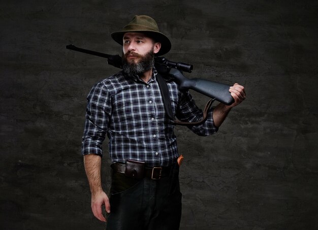 Bearded hunter in a fleece shirt and hat holds on his shoulder a rifle with sight, looking away. Isolated on a dark background.