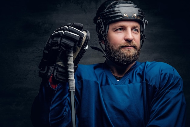 A bearded ice-hockey player in a blue sportswear holds a gaming stick in contrast illumination on grey vignette background.