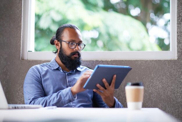 Bearded Indian man in glasses sitting at desk in office and using tablet