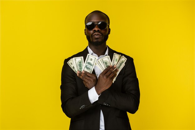 Bearded luxury young afroamerican guy is holding lots of money in both hands in sunglasses and black suit 