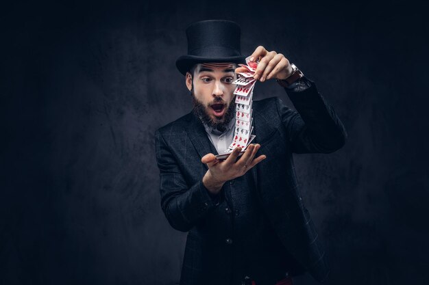 A Bearded magician in a black suit and top hat, showing trick with playing cards on a dark background.